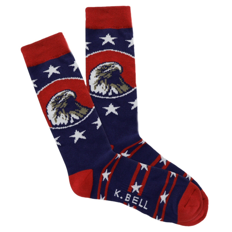 K Bell American Made Mens Bald Eagle Sock starts on a Navy background with Red Cuffs, Heels and Toes. The American Bald Eagle is in the center of a Red Oval in a Portrait. Around Him are Five Point Large White Stars in a Field of Blue. On the Foot are Blue Bars of smaller Five Point White Stars all around with Red small Stripes making each Bar.