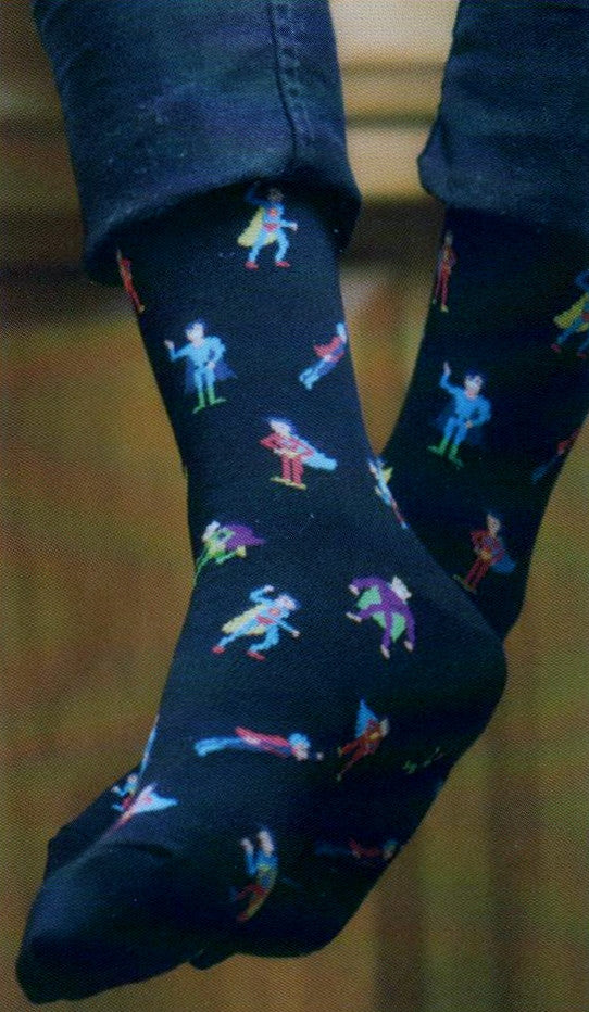 K Bell Mens Super Heroes Sock starts on a Black background. From the Cuffs to the Toes are Super Heroes in different colored outfits and capes. Some are flying, standing and running. Colors are Bright and pop off the Black in Red, Blue, Yellow. Green and Purple. This Sock is shown on a Model.