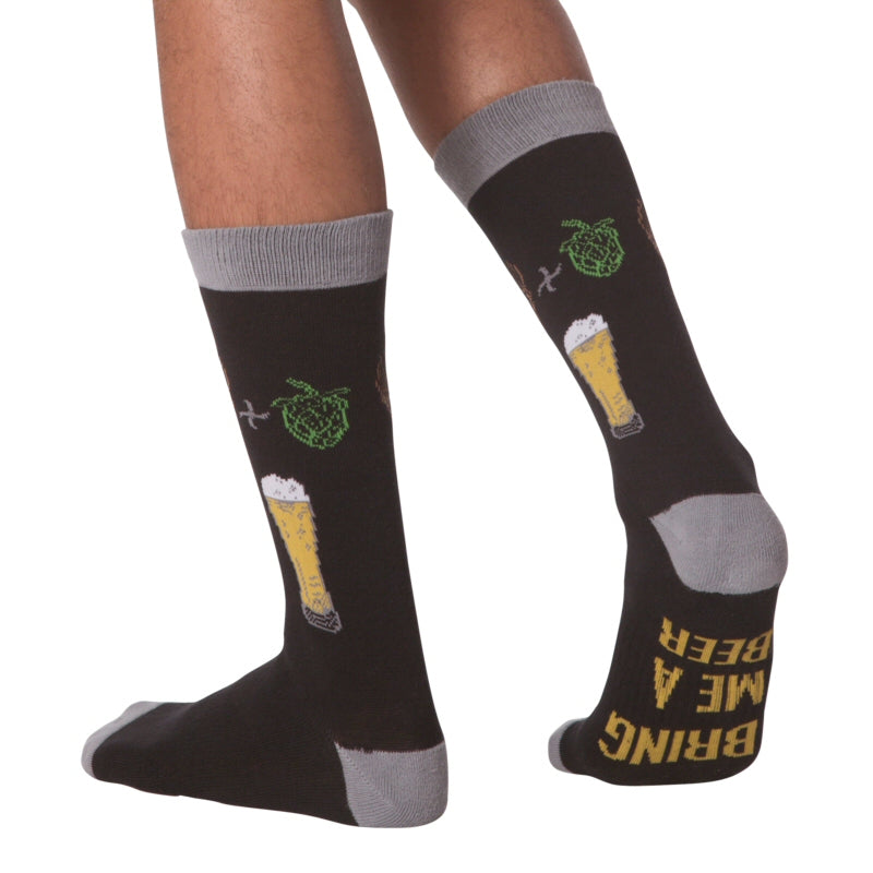 K Bell American Made Mens Beer Sock is shown Model. It starts on a Black background with Charcoal Grey Cuffs Heels and Toes. Under the Foot reads, "Bring Me A Beer" in Bold Golden Text. Below the Cuff are Brown Barley then a Plus Sign and Green Hops. Below is the Equals of a Glass holding a Beer, Golden with a Head of White Foam. 